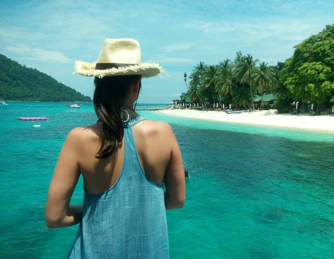 a woman in a hat looking out over the water at a beach and palm trees in the distance with a boat in the water